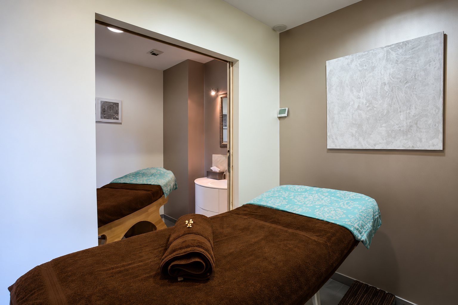 Une cabine de soin aux murs gris et taupe avec une table de massage couverte d'un drap brun et turquoise, et une seconde table visible dans la pièce voisine.