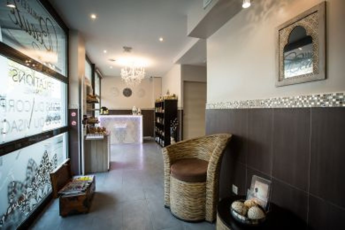 Couloir d'entrée d'un institut de beauté avec un fauteuil en osier, un lustre, des étagères de produits et une vitrine ornée de lettres.