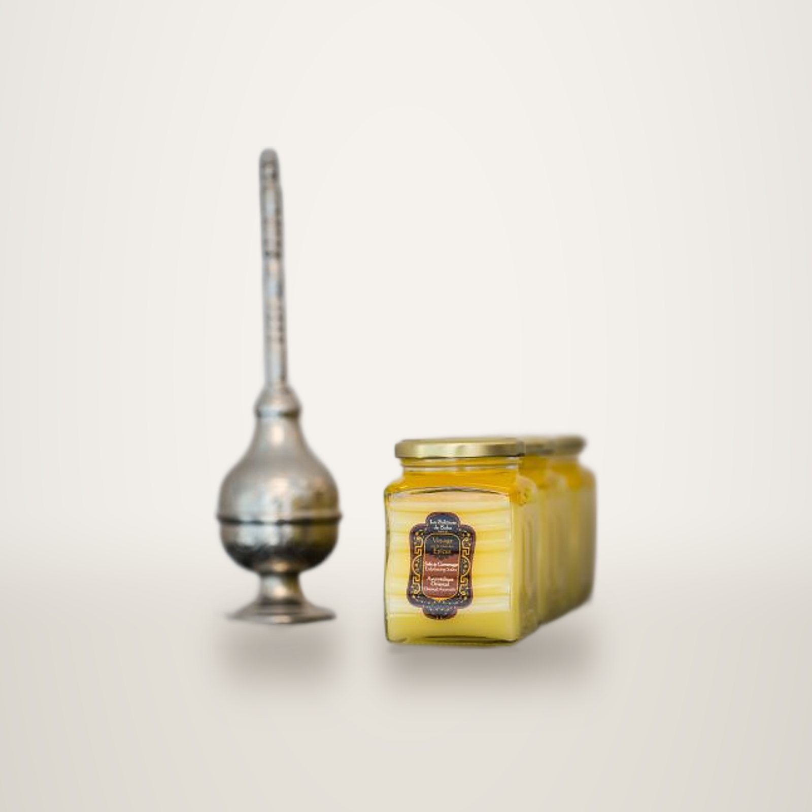 Des pots en verre remplis d'un baume jaune, avec une étiquette décorative, sont posés sur une étagère sombre à côté d'un flacon oriental en métal argenté.