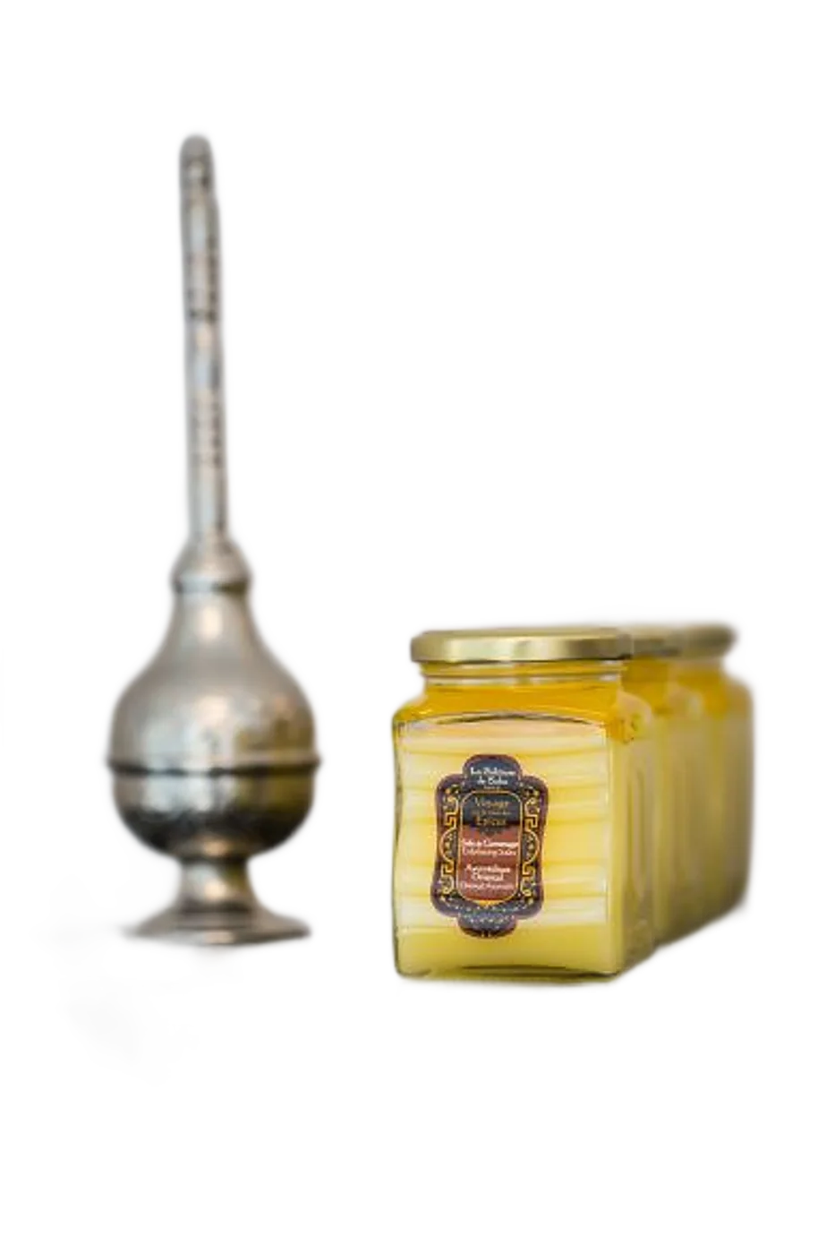 Des pots en verre remplis d'un baume jaune, avec une étiquette décorative, sont posés sur une étagère sombre à côté d'un flacon oriental en métal argenté.