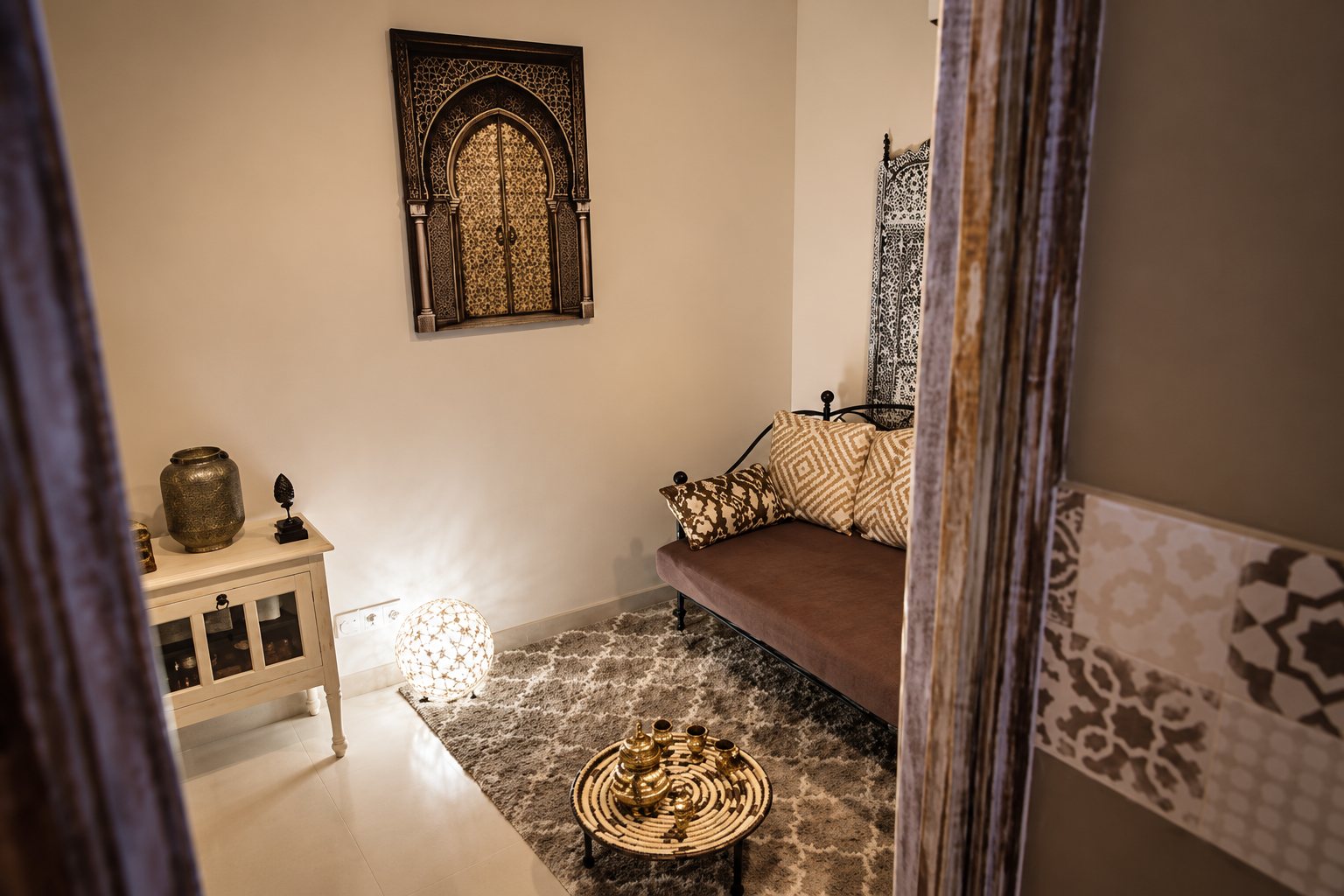 Petite pièce aux murs clairs décorée dans un style oriental, avec une banquette brune, des coussins, une lampe ajourée allumée, un plateau à thé sur le tapis et un cadre en forme d'arche au mur.
