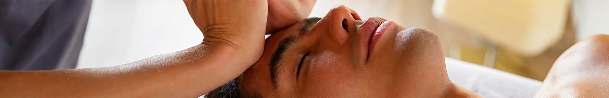 Une personne allongée les yeux fermés reçoit un massage du visage et de la tête, les mains d'un praticien posées sur son front.